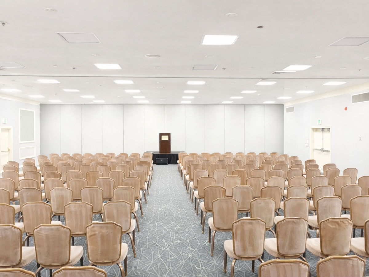 A large, empty conference room with rows of beige chairs facing a small stage and podium at the front.