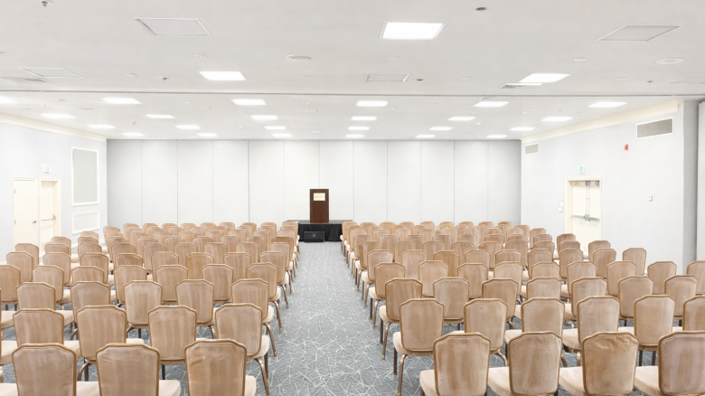 A large, empty conference room with rows of beige chairs facing a small stage and podium at the front.