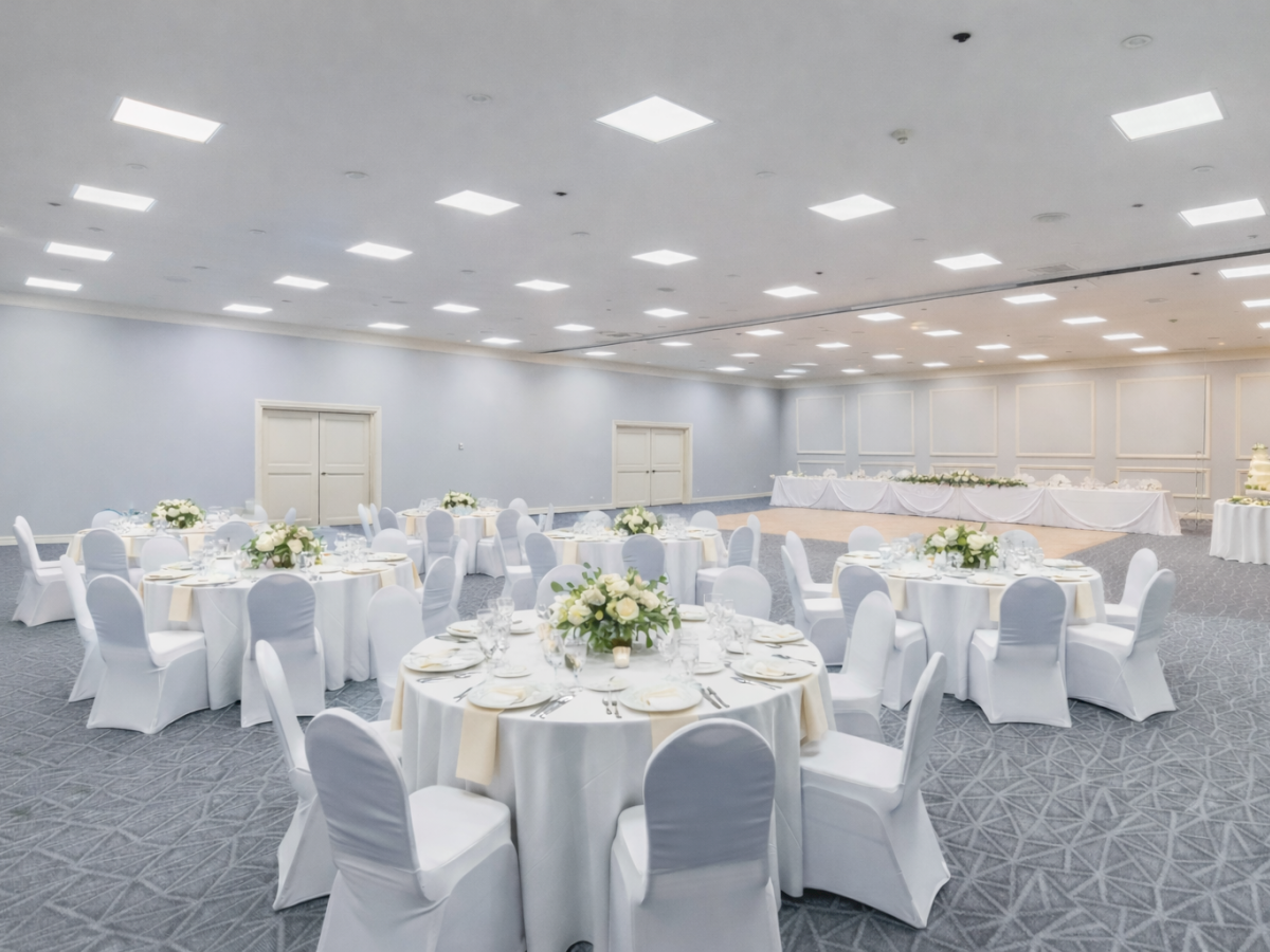 A banquet hall set for a formal event, with round tables draped in white cloth, floral centerpieces, and neatly arranged place settings.