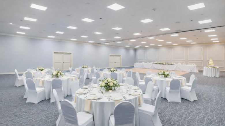 A banquet hall set for a formal event, with round tables draped in white cloth, floral centerpieces, and neatly arranged place settings.