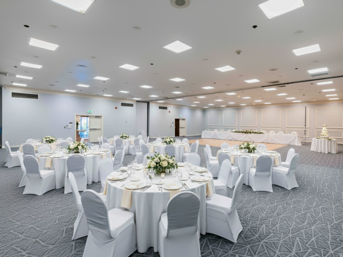A bright banquet hall with round tables, white chair covers, floral centerpieces, and a head table setup along a pale wall.