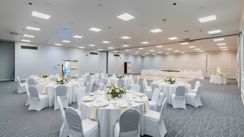 A bright banquet hall with round tables, white chair covers, floral centerpieces, and a head table setup along a pale wall.