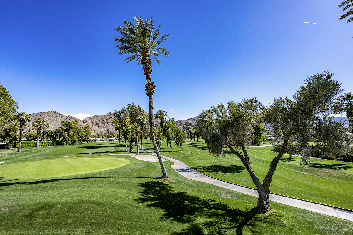 A sunny golf course with green fairways, palm trees, and a paved cart path winding through the landscape, mountains in the background.