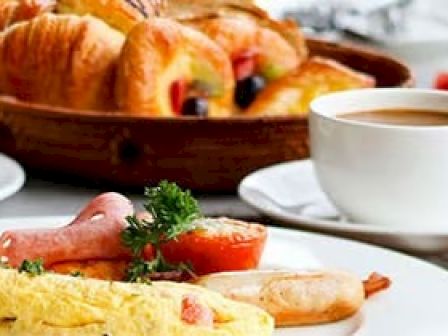 A breakfast plate with eggs, tomatoes, and sausage, plus croissants in the background and a cup of coffee on the side.