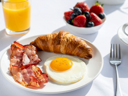 A breakfast plate with sunny-side-up egg, crispy bacon, a croissant, fresh berries, a glass of orange juice, and a cup of coffee.