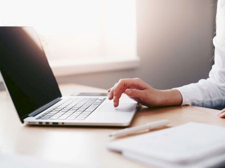 A person types on a laptop at a desk with papers nearby, bathed in natural light from a window.