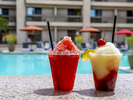 Two fruity shaved ice drinks by a pool, one red with a lemon wedge and ice, the other a layered yellow-red smoothie with a cherry on top.
