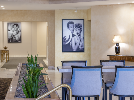 A stylish hotel lobby with beige walls, blue chairs, a water feature with plants, framed black-and-white photos, and a lit table lamp, inviting and calm.