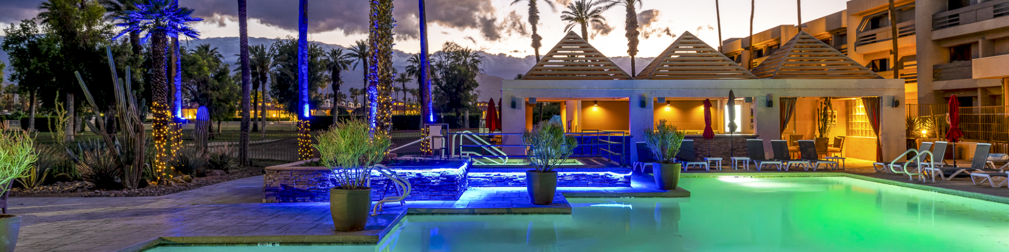 A resort-style pool area at dusk with palm trees, lounge chairs, and a lit building in the background, reflecting blue and green lights.