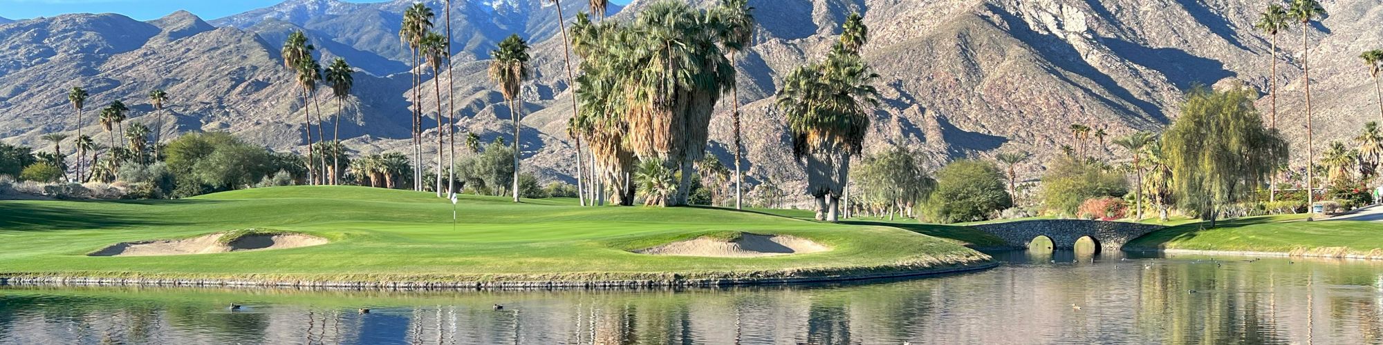 A serene golf course beside a calm lake, with palm trees, green fairways, and rugged mountains in the background under a clear blue sky.