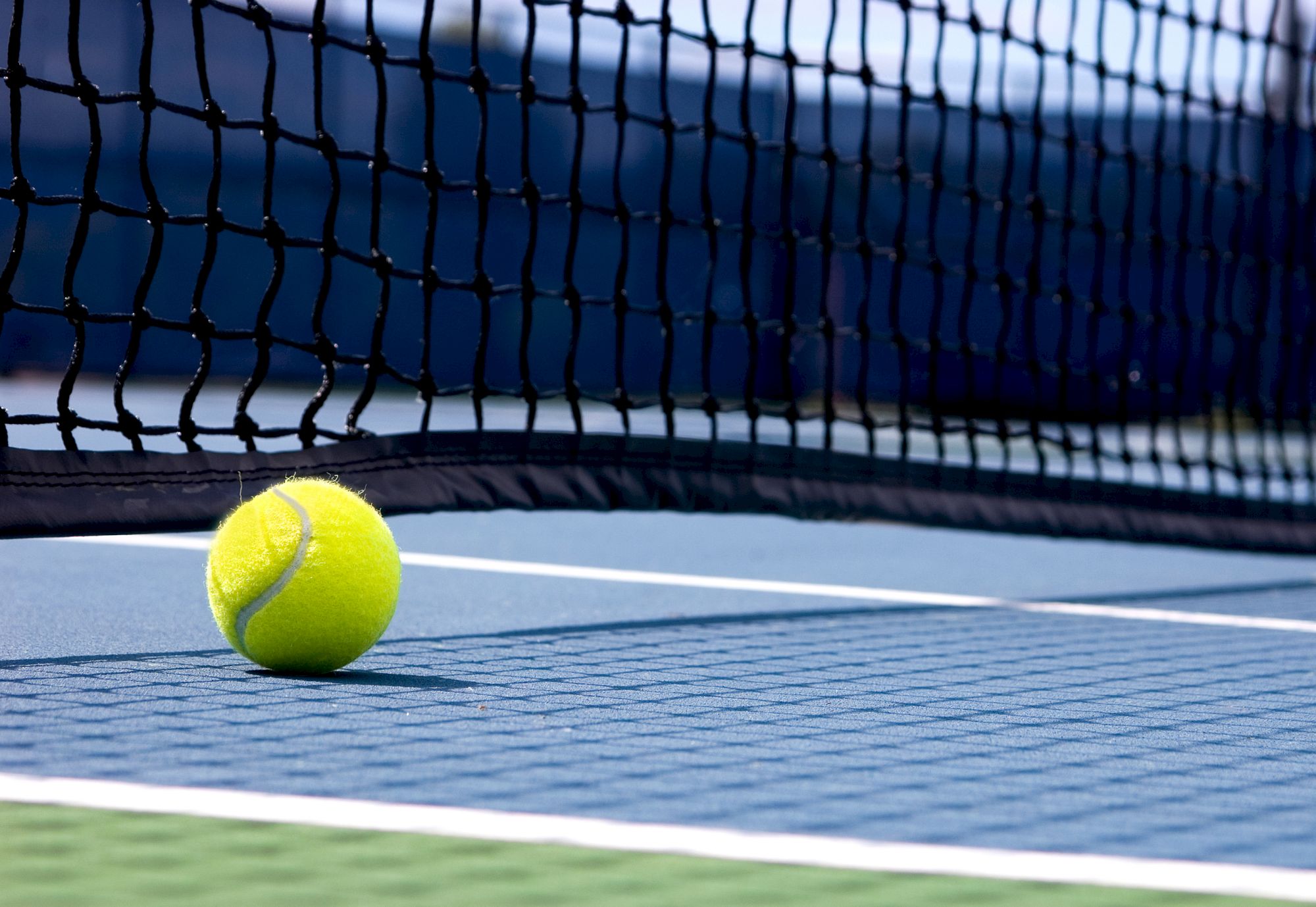 A yellow tennis ball rests near the net on a blue tennis court, with white boundary lines and a blurred background.