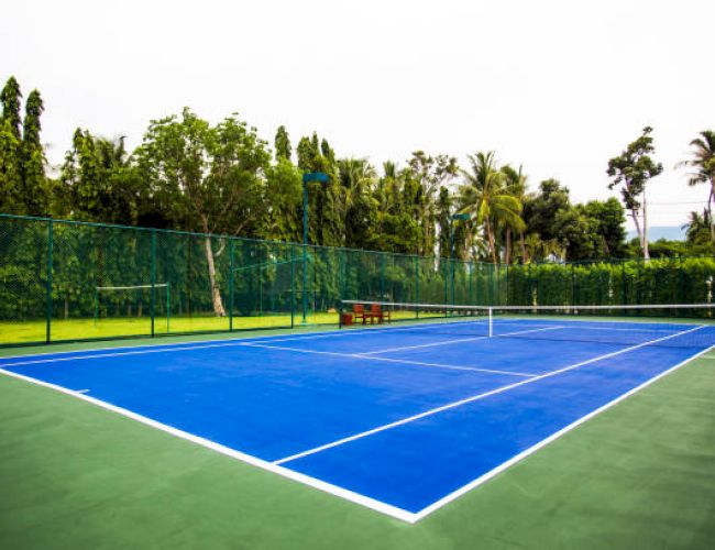 A bright blue tennis court with white lines, a net in the middle, surrounded by green fencing and lush trees in the background.