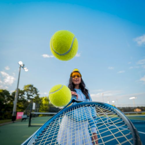 A girl in a headband tosses two large tennis balls toward the camera on a sunny tennis court, with a net in the foreground and blue sky.