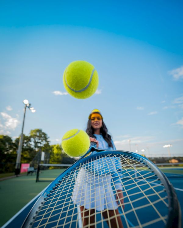 A girl in a headband tosses two large tennis balls toward the camera on a sunny tennis court, with a net in the foreground and blue sky.