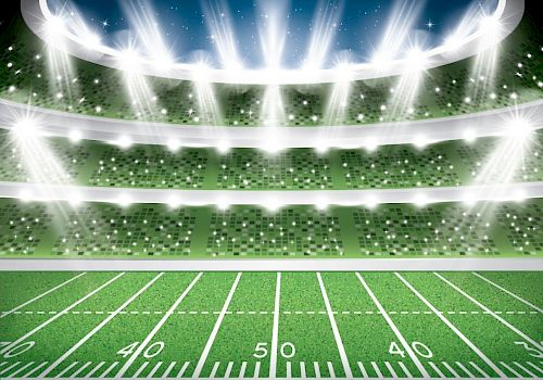 A bright football stadium at night with a lush green field and dramatic stadium lights blazing overhead.