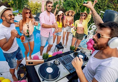 A DJ in headphones spins records at a poolside party, crowd dancing and cheering around him with bright summer vibes.