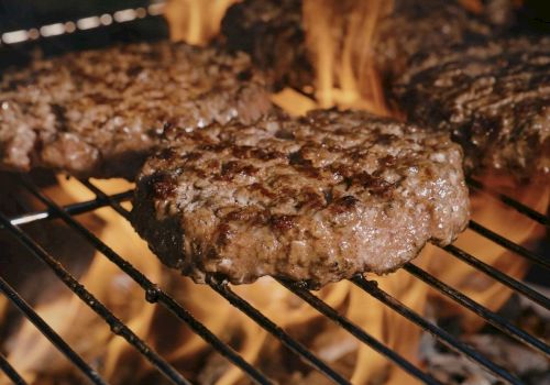 Juicy beef burgers grilling over an open flame, sizzling on the grates with charred edges and flames licking up around them.
