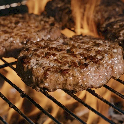 Juicy beef burgers grilling over an open flame, sizzling on the grates with charred edges and flames licking up around them.