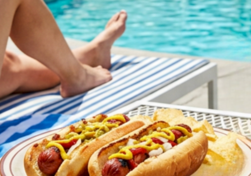 Two hot dogs loaded with toppings on a plate by the pool, with a margarita glass nearby and someone lounging in the background, sunny day.