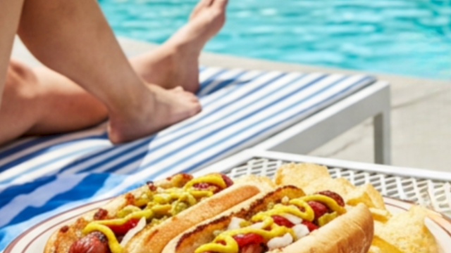 Two hot dogs loaded with toppings on a plate by the pool, with a margarita glass nearby and someone lounging in the background, sunny day.