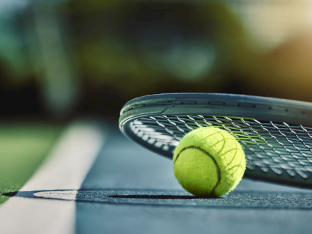 A tennis racket rests beside a bright tennis ball on a court, with a blurred green background and a white line beneath.