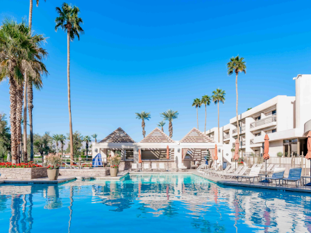 A sunny pool scene with blue water, palm trees, lounge chairs, and a resort building in the background.