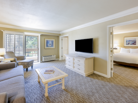 A spacious hotel suite with a seating area, coffee table, TV on a dresser, and a doorway to a separate bedroom; neutral tones throughout.