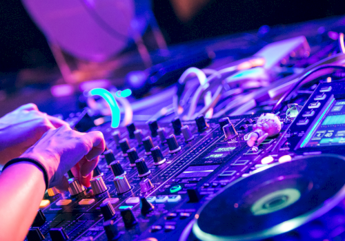 A DJ&rsquo;s hands on a colorful mixer, knobs, lights, and turntables in a vibrant club setup, creating a lively electronic music vibe.