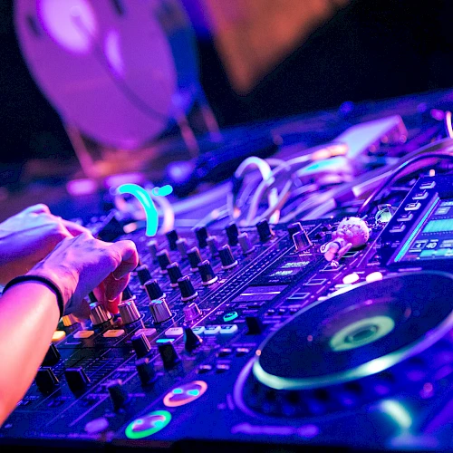 A DJ&rsquo;s hands on a colorful mixer, knobs, lights, and turntables in a vibrant club setup, creating a lively electronic music vibe.