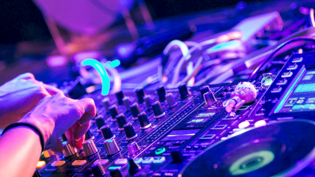 A DJ&rsquo;s hands on a colorful mixer, knobs, lights, and turntables in a vibrant club setup, creating a lively electronic music vibe.
