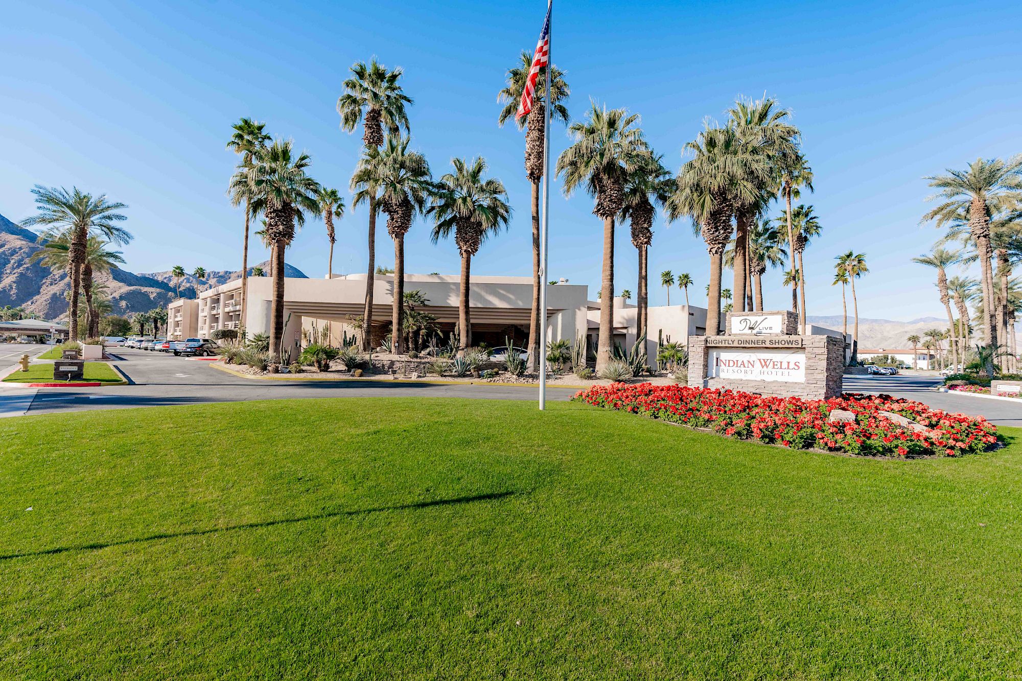 A sunny scene with palm trees, a grassy lawn, a roadside sign, and a low building complex against a clear blue sky.