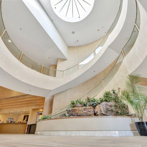 A modern, spiraling lobby interior with white curved walls, a central skylight, and greenery on a stepped planter near a curved staircase.
