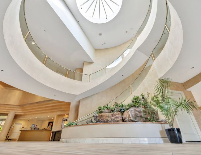 A modern, spiraling lobby interior with white curved walls, a central skylight, and greenery on a stepped planter near a curved staircase.