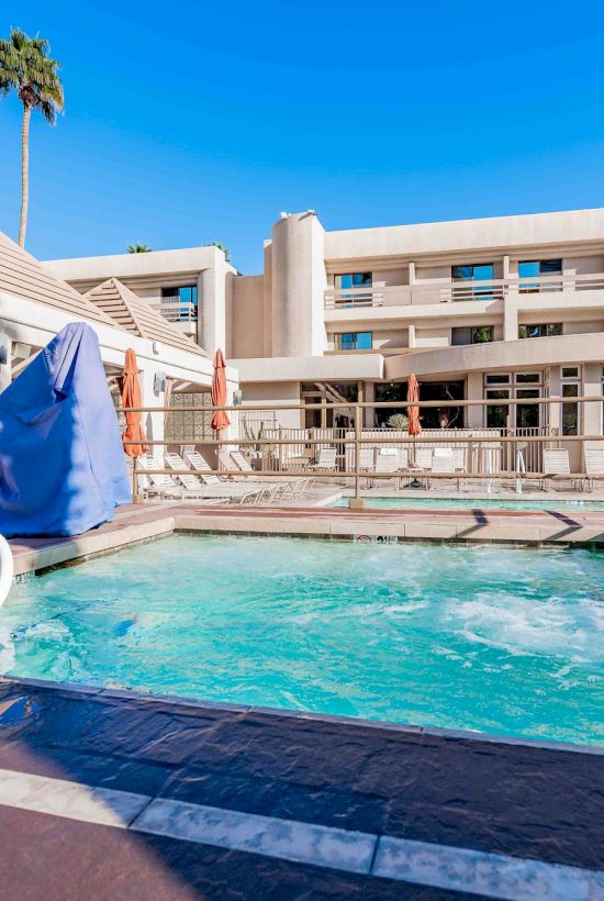 A hotel pool area with a bright blue sky, pool, lounge chairs, and a multi-story building in the background.
