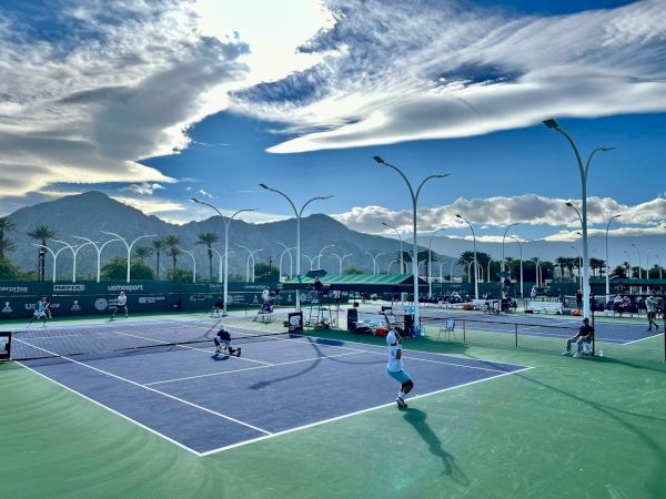 A sunny outdoor tennis court with players in action, modern lighting poles, and mountains in the background under a dramatic blue sky.