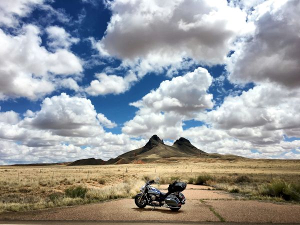 A motorcycle parked on a cracked road in a wide, arid landscape with distant hills and dramatic, fluffy clouds overhead.