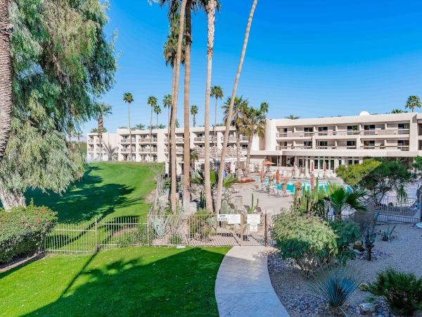 A sunny resort courtyard with green lawns, palm trees, a pool area, and a low-rise beige hotel building in the background. End.