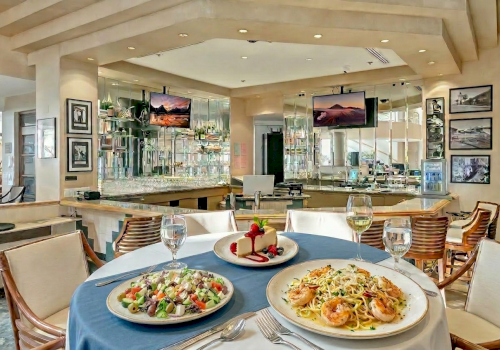 A stylish restaurant dining area with a round table of pasta and salad, wine glasses, and a scenic bar in the background, inviting and airy.