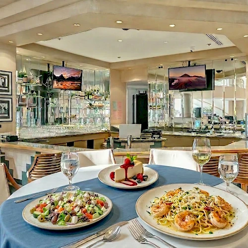 A stylish restaurant dining area with a round table of pasta and salad, wine glasses, and a scenic bar in the background, inviting and airy.