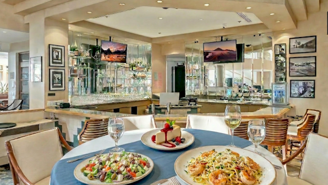 A stylish restaurant dining area with a round table of pasta and salad, wine glasses, and a scenic bar in the background, inviting and airy.