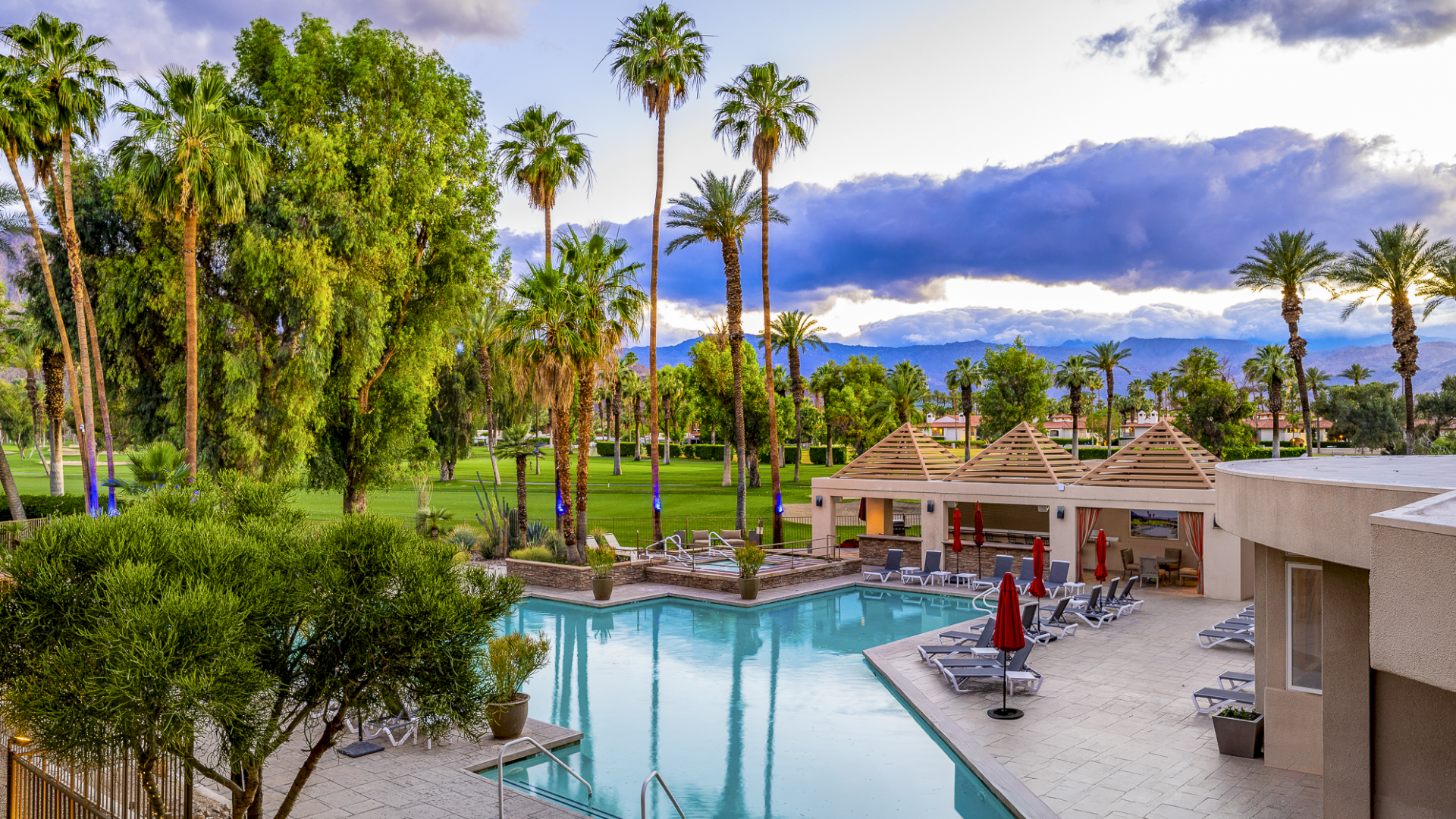 A resort-style pool area with clear blue water, loungers, palm trees, and small cabana-style buildings under a partly cloudy sky. The scene feels relaxing and tropical.