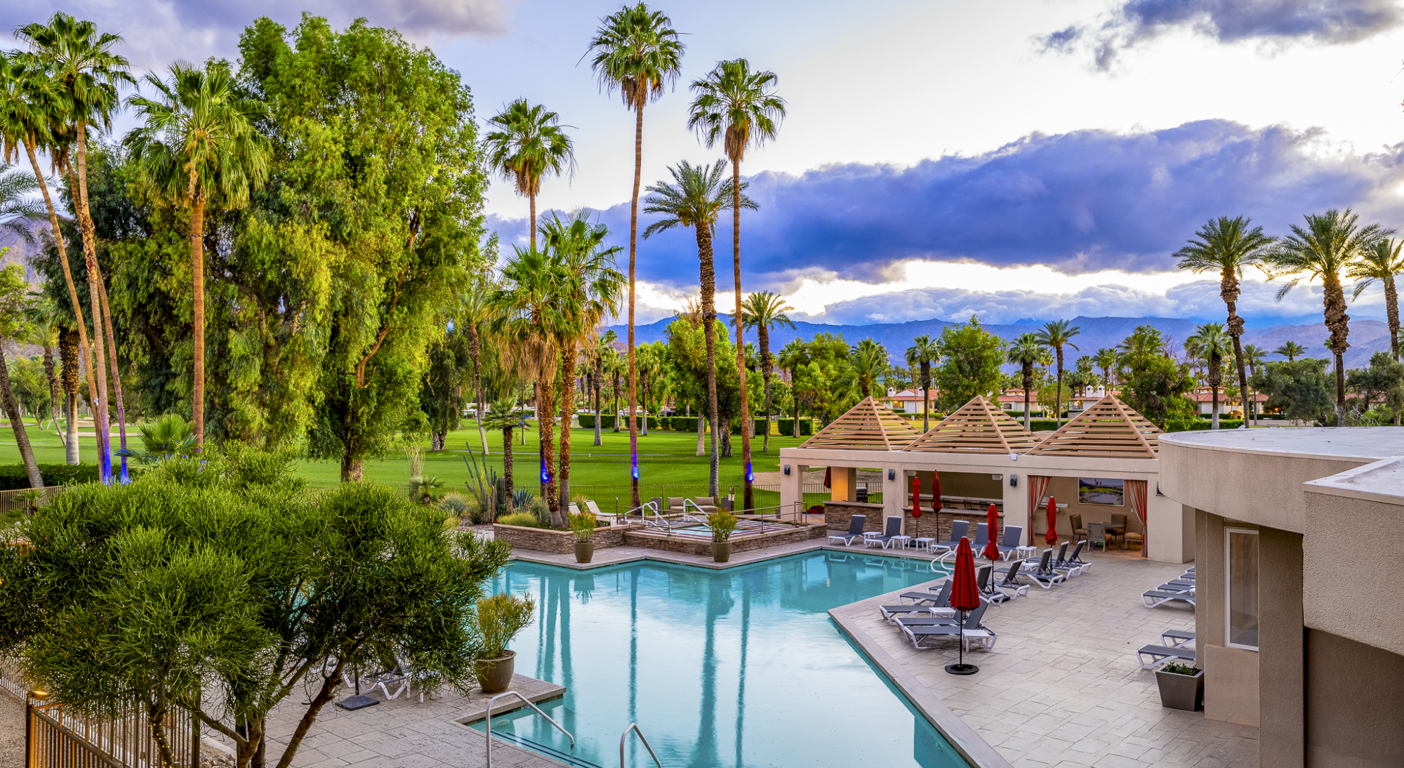 A resort-style pool area with clear blue water, loungers, palm trees, and small cabana-style buildings under a partly cloudy sky. The scene feels relaxing and tropical.