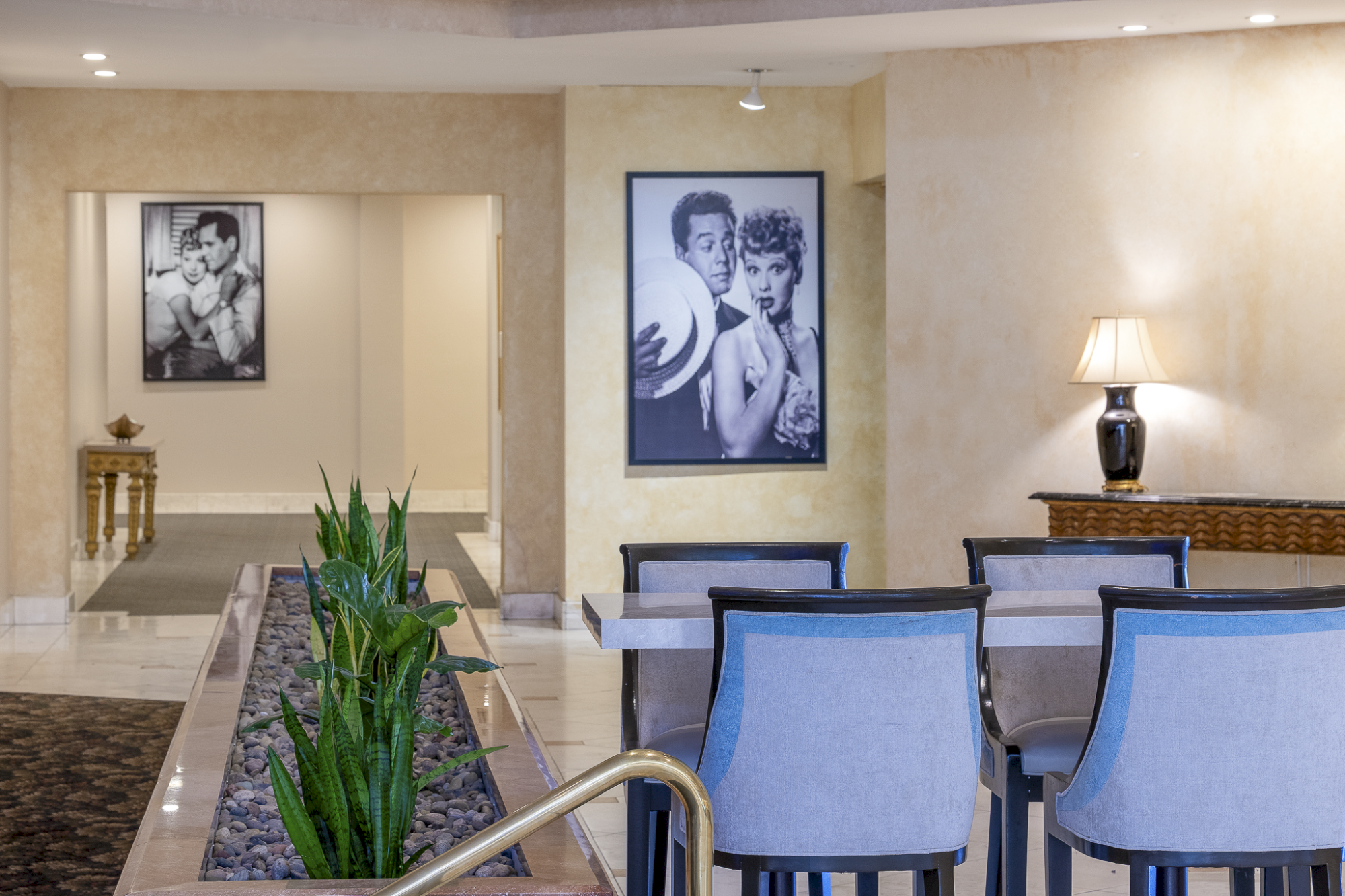 A chic hotel lobby with beige walls, blue-upholstered chairs, a central planter, black-and-white framed photos, and a lit table lamp, calm and upscale.