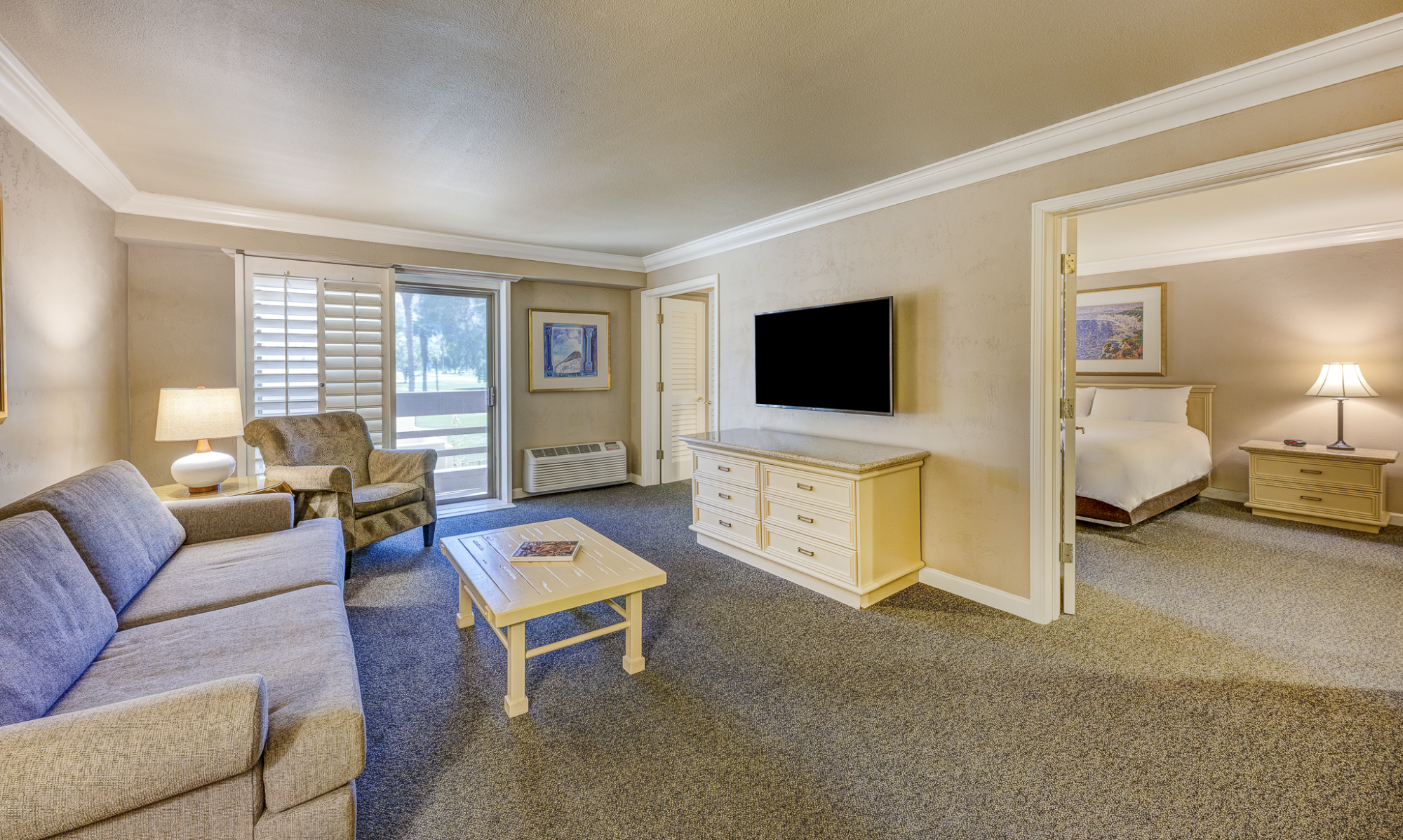 A spacious hotel suite living area with a gray sofa, armchairs, coffee table, TV on a dresser, and a doorway to a bedroom, all softly lit.