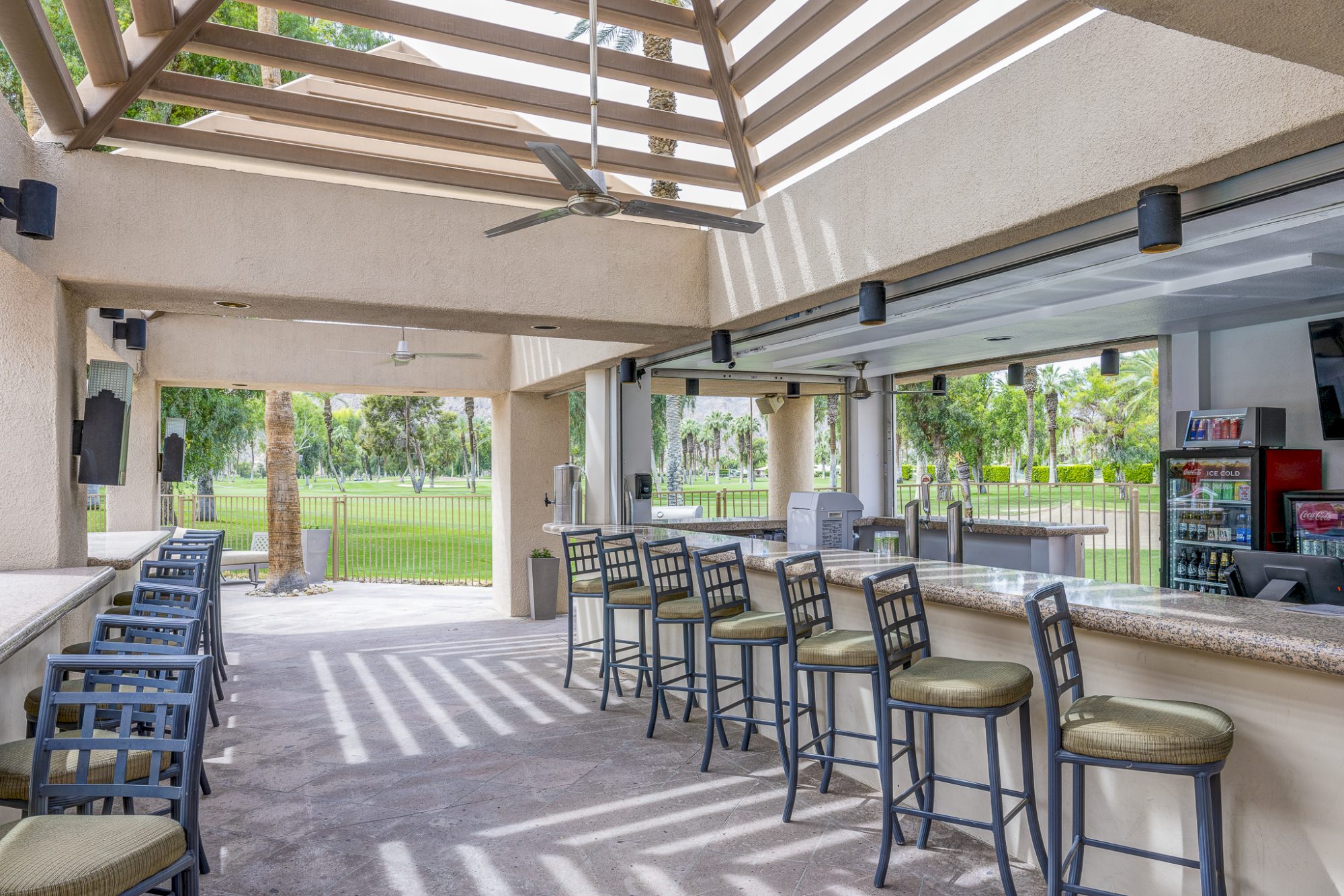 A bright outdoor bar area with a long counter, bar stools, ceiling fans, shaded rafters, and a view of a green field beyond.