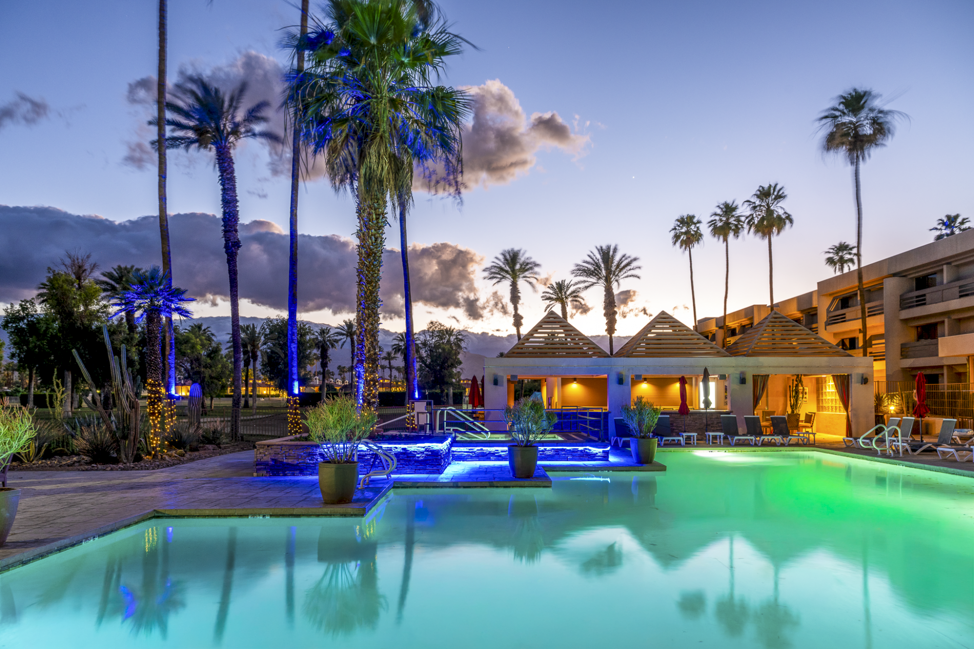 A resort pool at dusk with palm trees, glowing waters, lounge areas, and a modern building backdrop, inviting and tranquil.