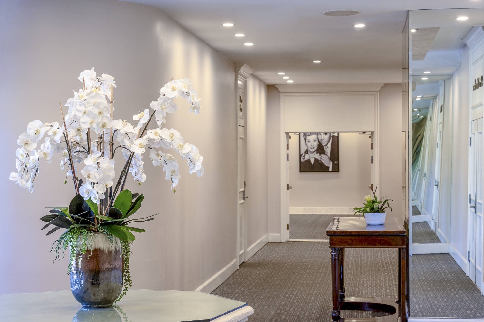 A bright hallway with white walls, a wooden table, and a large vase of white orchids on a round table in the foreground, plus framed art and plants.