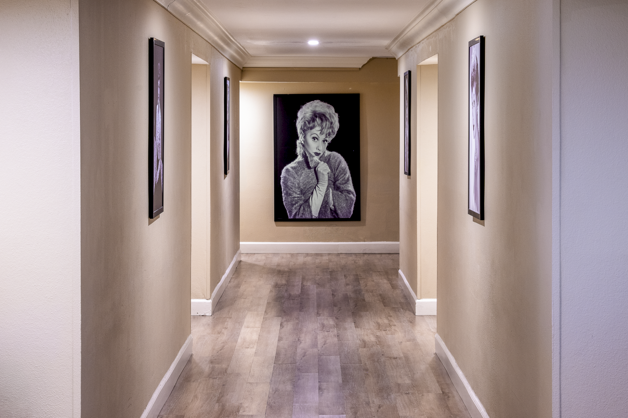A hallway with beige walls, framed portraits along the sides, and a large monochrome portrait at the far end, hallway lighting above.
