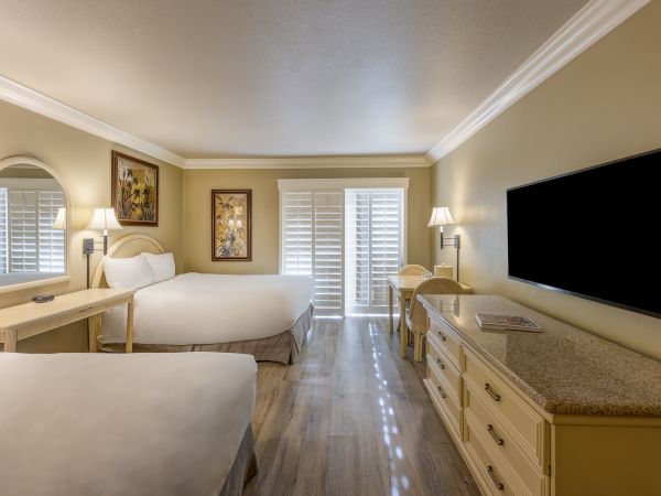 Two twin beds in a bright hotel room with a desk, wall mirror, dresser, and a mounted TV; glass doors with shutters at the far end.