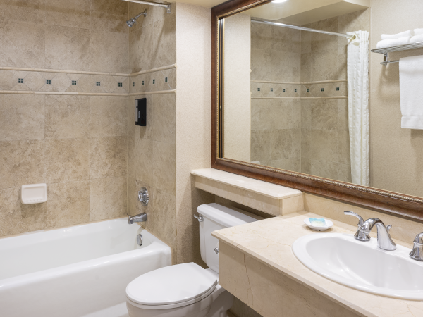 A beige-tiled bathroom with a bathtub, toilet, and double-sink vanity; a large mirror above the sink and a towel rack. End with a period.